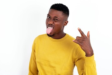 Portrait of a crazy young handsome man wearing yellow sweater over white background showing tongue horns up gesture, expressing excitement of being on concert of band.