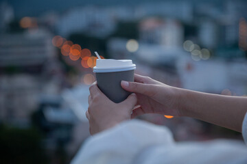 A woman's hand holding a cup of coffee or tea with a view with buildings in the evening light and inspiring beautiful bokeh lights on an abstract city background at sunrise or sunset. Soft spot focus