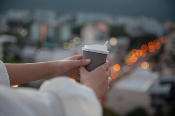 A woman's hand holding a cup of coffee or tea with a view with buildings in the evening light and...