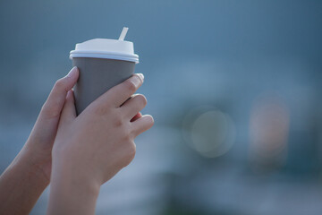 A woman's hand holding a cup of coffee or tea with a view with buildings in the evening light and...