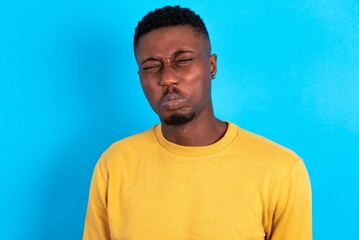 Fototapeta premium Dismal gloomy rejected young handsome man wearing yellow T-shirt over blue background has problems and difficulties, curves lower lip and closes eyes in despair, being in depression