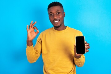 young handsome man wearing yellow T-shirt over blue background holding in hands cell showing ok-sign