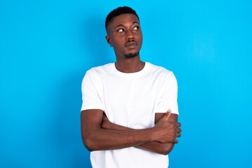 Fototapeta premium Pleased young handsome man wearing white T-shirt over blue background keeps hands crossed over chest looks happily aside