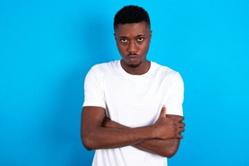 Gloomy dissatisfied young handsome man wearing white T-shirt over blue background looks with miserable expression at camera from under forehead, makes unhappy grimace