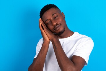 young handsome man wearing white T-shirt over blue background leans on pressed palms closes eyes and has pleasant smile dreams about something