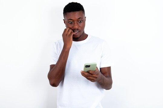 Portrait of pretty frightened young handsome man wearing white T-shirt over white background chatting biting nails after reading some scary news on her smartphone.