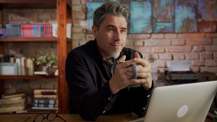 Smiling man using laptop on table at home, browsing internet, working in home office drinking morning coffee.