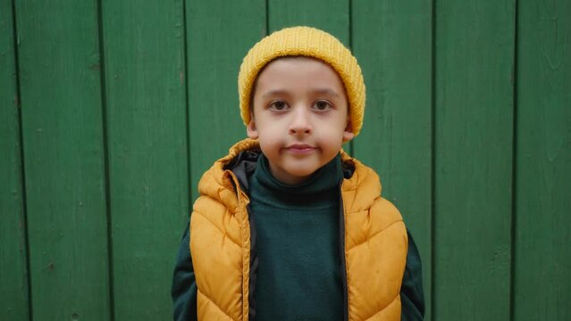 a boy child in a yellow vest in a knitted hat stands against the background of a green wooden fence in the village. Maple leaves fall from the sky on the boy
