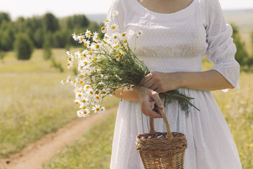 Faceless woman in vintage white dress walking along a rural road with a bouquet of daisies and a...