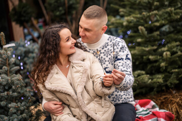 Fototapeta premium A beautiful young couple sitting near Christmas trees in white clothes walking near garlands. Happy man and woman, romance, Christmas celebration, fun, love. White sweater, new year, street, outside