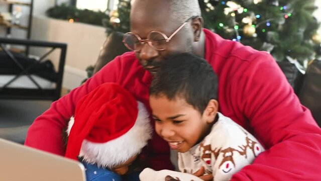dad, son and daughter in christmas sweaters and santa hat hug, smile and communicate via video link with relatives on the background of the christmas tree.