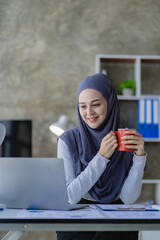 A beautiful Asian Muslim woman wears a headscarf using her laptop to do financial accounting work in her living room at home.