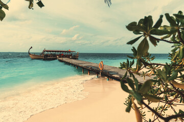Uitzicht op een houten pier die leidt naar een prachtige gondel die aan het einde ervan is afgemeerd, omgeven door blauwgroen oceaanwater  koraalzand van een Maldivisch eiland met groen en een golfschuim op de voorgrond