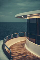 Fototapeta premium A vertical low-key view of a diving safari yacht's middle deck with a chrome fence, lit wooden floor, illuminated ceiling, and windows reflecting the dark ocean waterscape and part of the evening sky