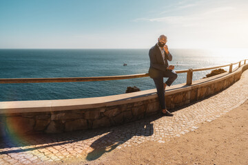 A bald stylish African-American man with a black beard and in an elegantly tailored summer costume is leaning against shoreline fencing, with an ocean behind and a copy space area on the left