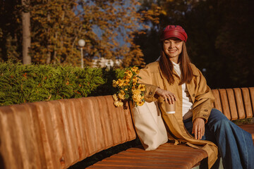 Positive young woman in trench with paper cup of coffee to go sitting on bench with flowers in bag and looking at camera while enjoying sunny day in autumn park 