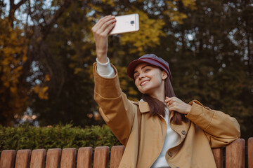 Low angle of young positive woman in trench and cap taking self portrait on cellular phone while having fun in park in fall season 