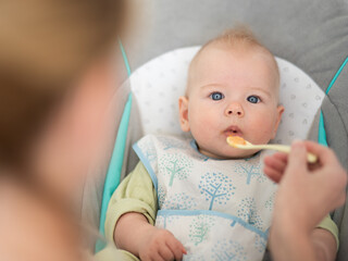 Mother spoon feeding her baby boy infant child in baby chair with fruit puree. Baby solid food introduction concept.