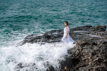A woman stands on a rock in the sea during a storm. Dressed in a white long dress, the waves break on the rocks and white spray rises.