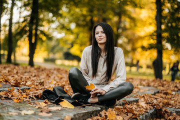 Young beautiful woman with dark hair in a leather jacket walks in the park in autumn