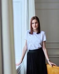 A young stylish girl is standing at the window in a bright room. Style and fashion. A young model.