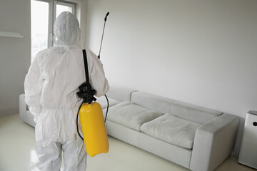 Worker in white uniform with sprayer standing in apartment