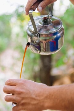 Vertical closeup of hands holding a cup and pouring coffee into it from a metallic kittle