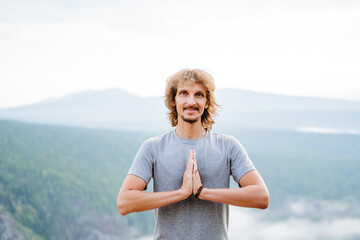 Fototapeta na wymiar The guy makes a bow greeting in the oriental style, the man folded his hands in front of his chest, namaste, smile, long hair, a young man in the mountains meditates,