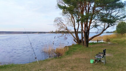 On the shore of the lake there is a place for fishing. There is a chair, on stands are installed feeder rods with winchless reels, there is a net, a bucket with bait and a box with baits