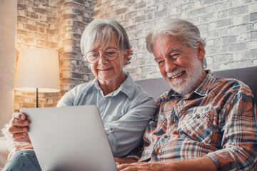 Cute couple of old people sitting on the sofa using laptop together shopping and surfing the net. Two mature people in the living room enjoying technology.