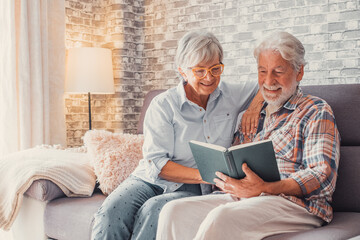 Portrait of couple of two cute grandparents reading a book together sitting on the sofa. Grandmother and grandfather relaxing weekend on free time.