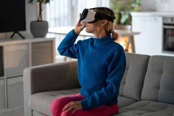 Fototapeta premium Resting woman freelancer wearing VR glasses enjoys spending weekend playing futuristic games in apartment. Young female wearing blue sweater sits on comfortable sofa and explores virtual world gaming