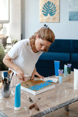 Woman artist painting picture at home with acrylic paints and spatula over the window. Adult hobby and free time creative activity. Female at her 40s enjoying art. 