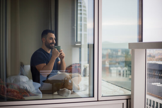 Smiling young adult man holding a cup of coffee, sitting on a balcony, a man resting or taking a break from hard work