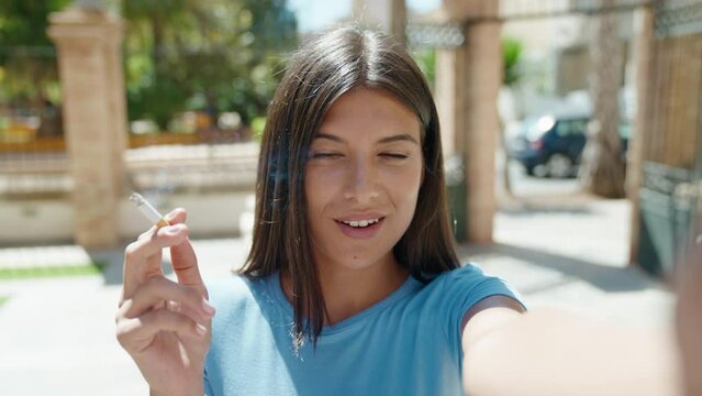Young beautiful hispanic woman having video call smoking at street