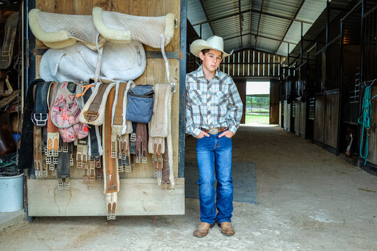 Young cowboy standing in a horse barn with saddles and other riding equipment stowed nearby