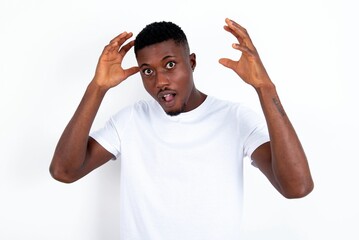 Fototapeta premium young handsome man wearing white T-shirt over white background looks with excitement at camera, keeps hands raised over head, notices something unexpected, reacts on sudden news.