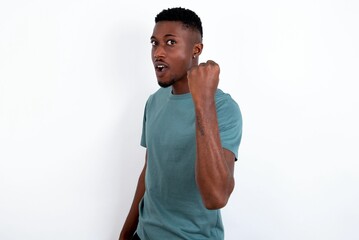 Overjoyed young handsome man wearing green T-shirt over white background glad to receive good news, clenching fist and making winning gesture.