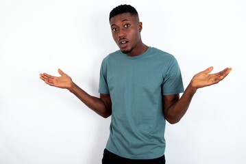 So what? Portrait of arrogant young handsome man wearing green T-shirt over white background shrugging hands sideways smiling gasping indifferent, telling something obvious.