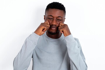 Pleased young handsome man wearing grey sweater over white background with closed eyes keeps hands near cheeks and smiles tenderly imagines something very pleasant