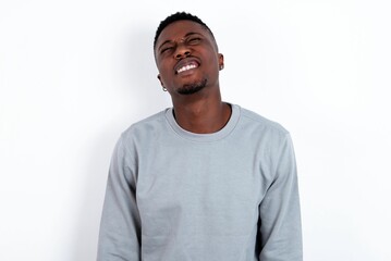 Positive young handsome man wearing grey sweater over white background with overjoyed expression closes eyes and laughs shows white perfect teeth