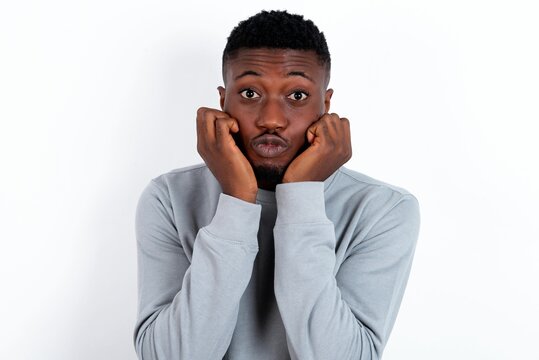 young handsome man wearing grey sweater over white background with surprised expression keeps hands under chin keeps lips folded makes funny grimace