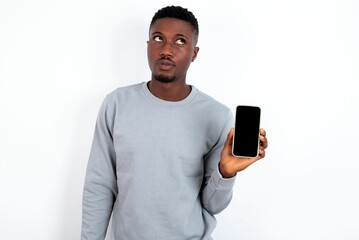 young handsome man wearing grey sweater over white background holds new mobile phone and looks mysterious aside shows blank display of modern cellular
