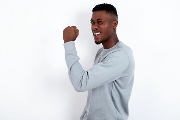 Portrait of funny young handsome man wearing grey sweater over white background shout yeah raise fists hands celebrate victory game competition