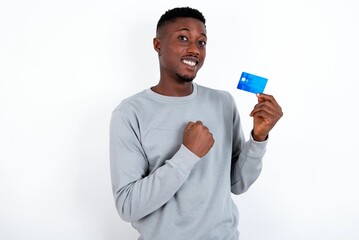 Excited happy positive cheerful smiling young handsome man wearing grey sweater over white background hold credit card raise fist in victory