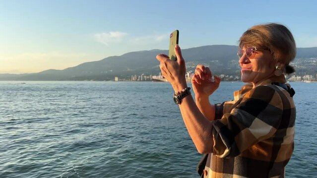 adult woman in sunglasses in a plaid brown jacket A woman in her hands with a phone and a watch on her hand shoots a video on the iPhone Stanley Park Vancouver Canada with mountains in the background