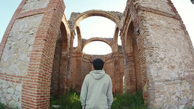 Young man stops in front of an old ruined chapel architecture and walks in. Spain.