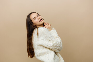 a beautiful, sweet, relaxed, attractive woman stands sideways in a white sweater, posing on a beige background, raising her arms bent at the elbows to her face and looks into the camera