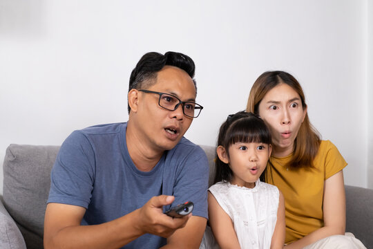 Asian Mother, Father and their daughter sit on sofa couch and feeling excited and happy wile watching the movie from television inside of the living room. Family activity during weekend.