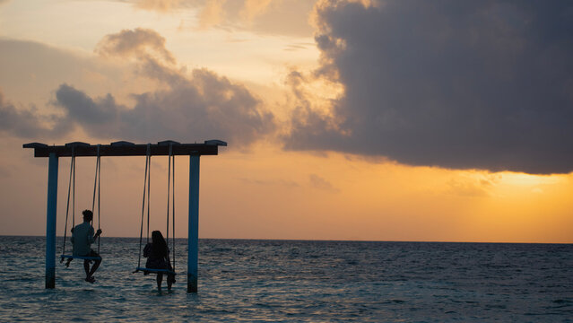 A picture of two couples sitting on a swing in the shallow sea of madives and looking at the beautiful orange sky during sunset